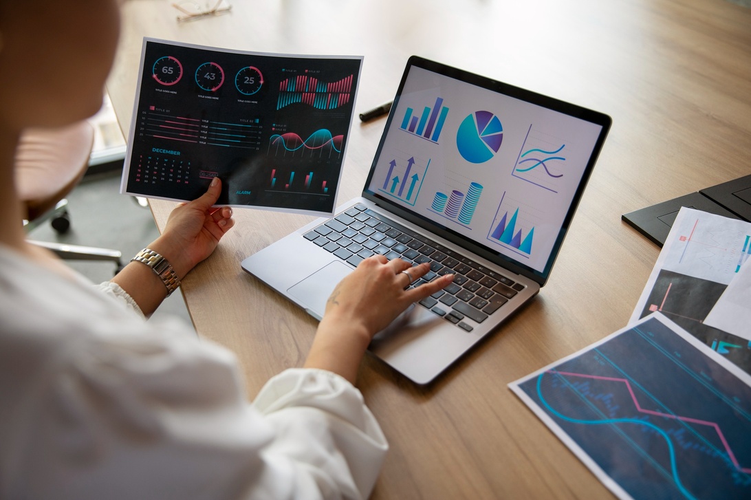 En person som analyserar affärsdata och diagram på både en bärbar dator och en separat skärm eller surfplatta. Olika färgglada grafer och diagram är synliga på båda skärmarna.