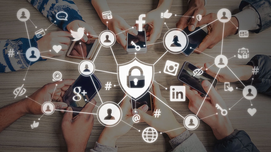 Flera händer som håller smartphones på ett träbord, med sociala medier-ikoner och nätverkslinjer överlagda, samt en central säkerhetsikon. Detta representerar övervakning och datainsamling från sociala nätverk.