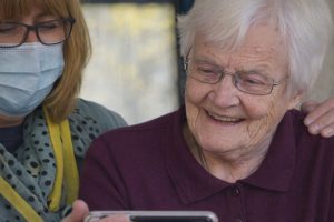 En äldre kvinna ler när hon tittar på en mobilskärm tillsammans med en person i munskydd.